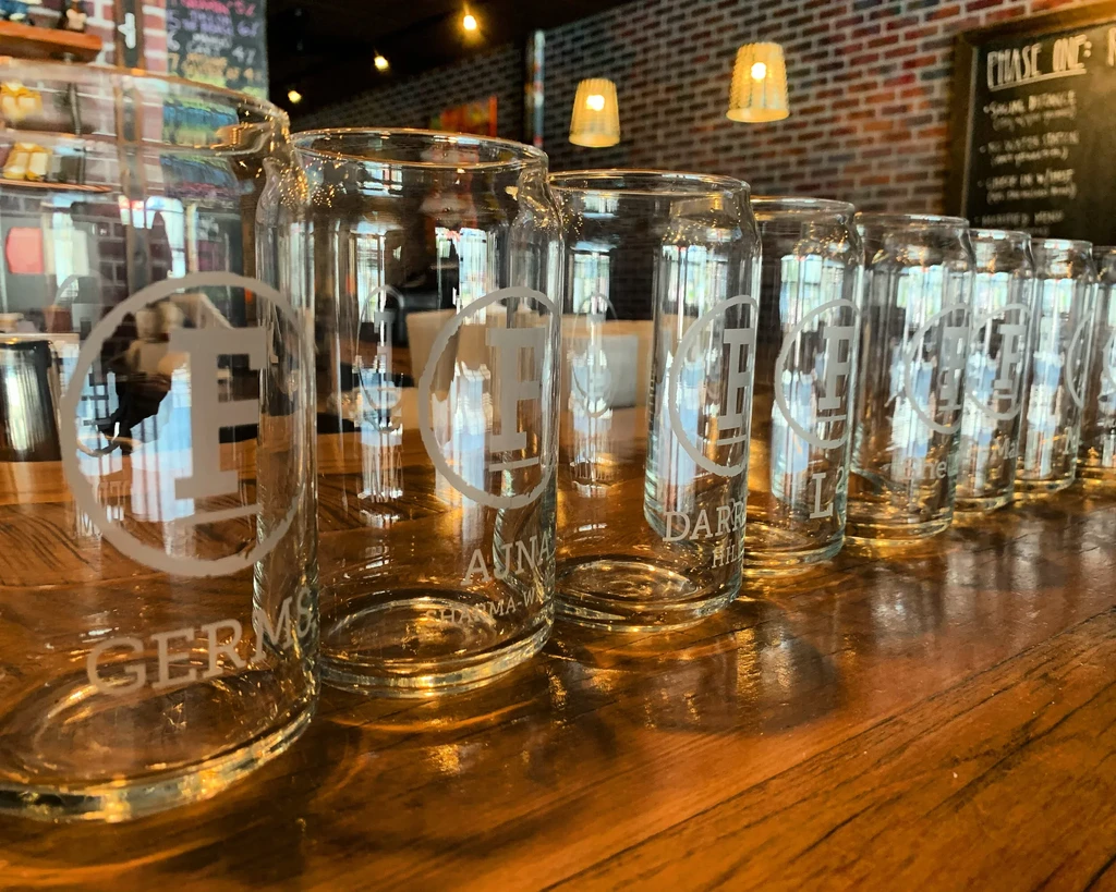 Empty can-shaped glasses with etched logos line a wooden bar.