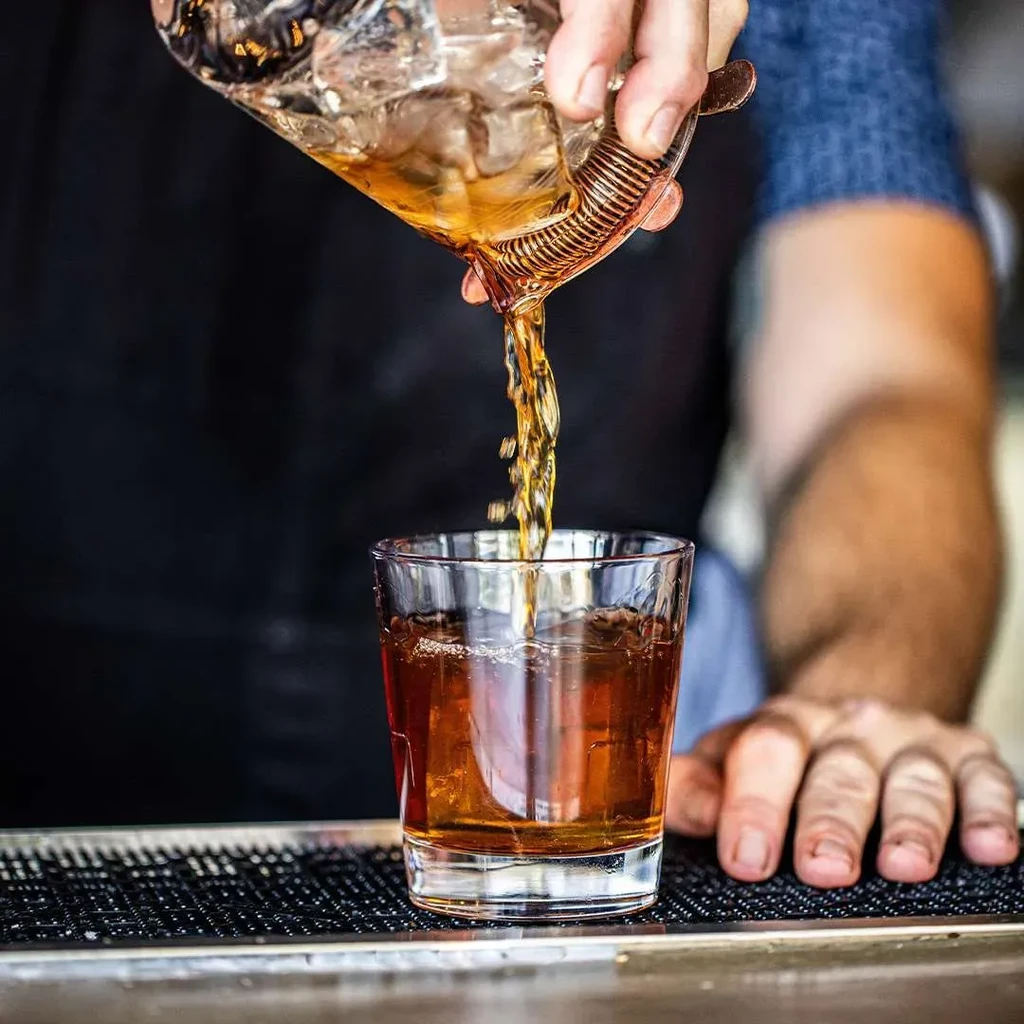 Golden liquid pours from mixing glass through strainer into rocks glass.