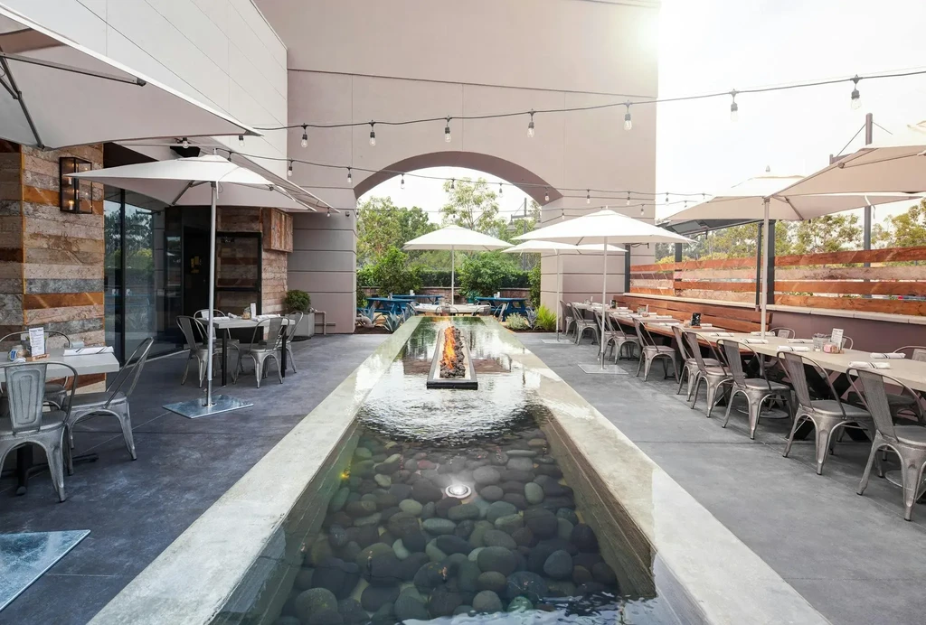 Modern outdoor dining patio with water feature, fire pit, tables, umbrellas.