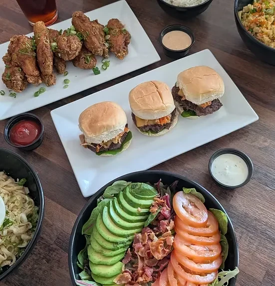 Overhead view of fried chicken, sliders, avocado salad, and noodles on wood table.