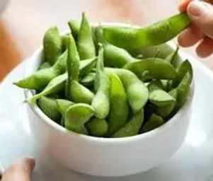 Hand picking a bright green edamame pod from a white bowl.