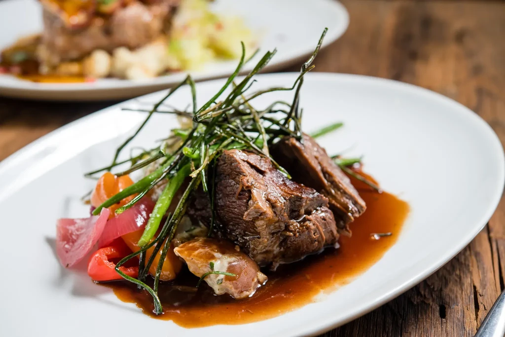 Plated meat, dark sauce, crispy herbs, colorful vegetables on white plate.