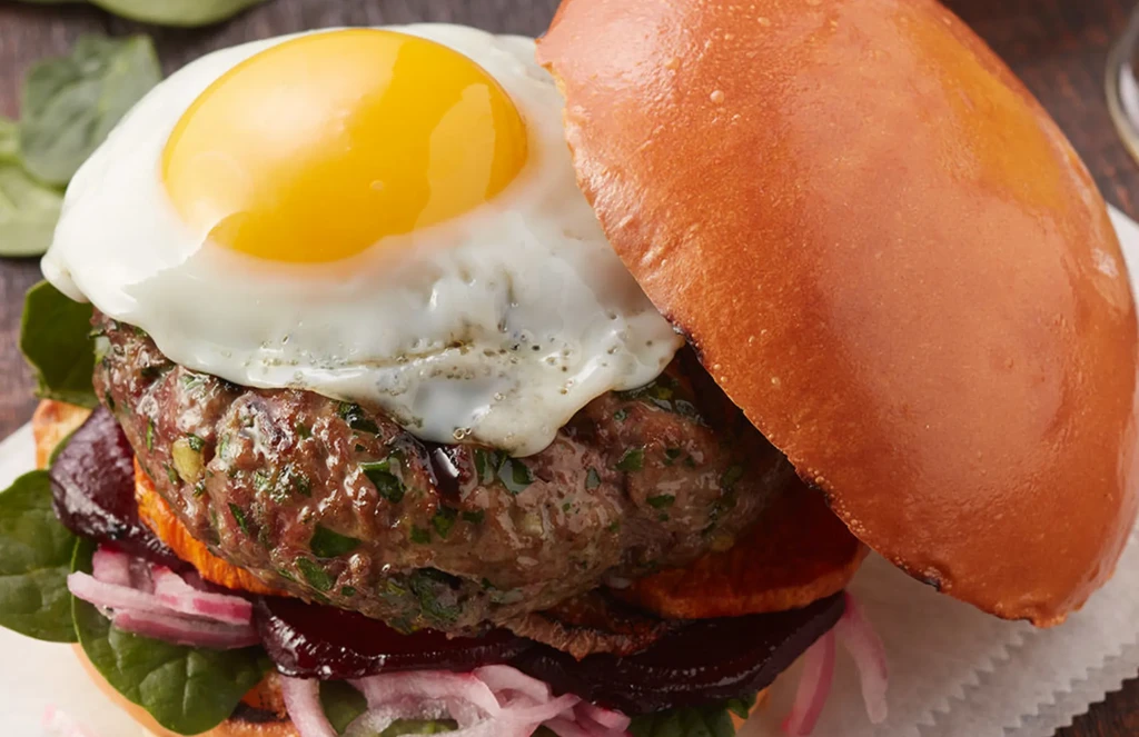 Stacked gourmet burger with fried egg, beef patty, beets, and spinach.