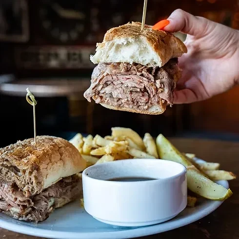 A hand holds a French dip sandwich half, with fries and au jus.