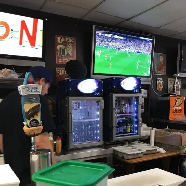 Bartender, beer tap, fridges, and TVs showing soccer in a casual bar.