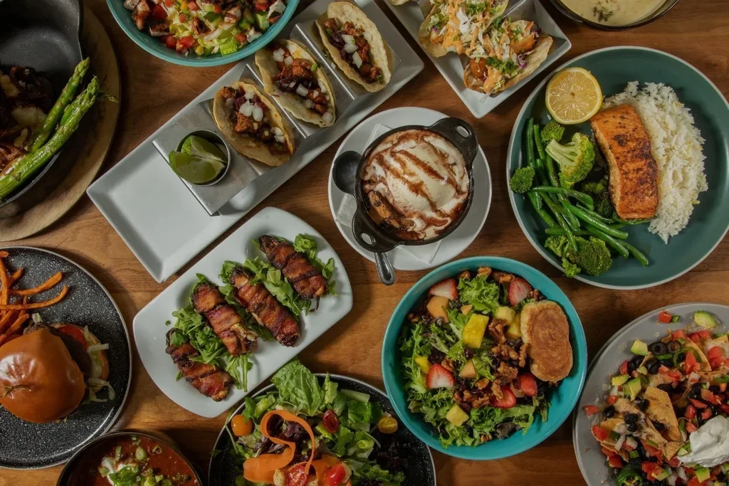 Diverse food spread on a wooden table: tacos, salmon, burger, dessert.