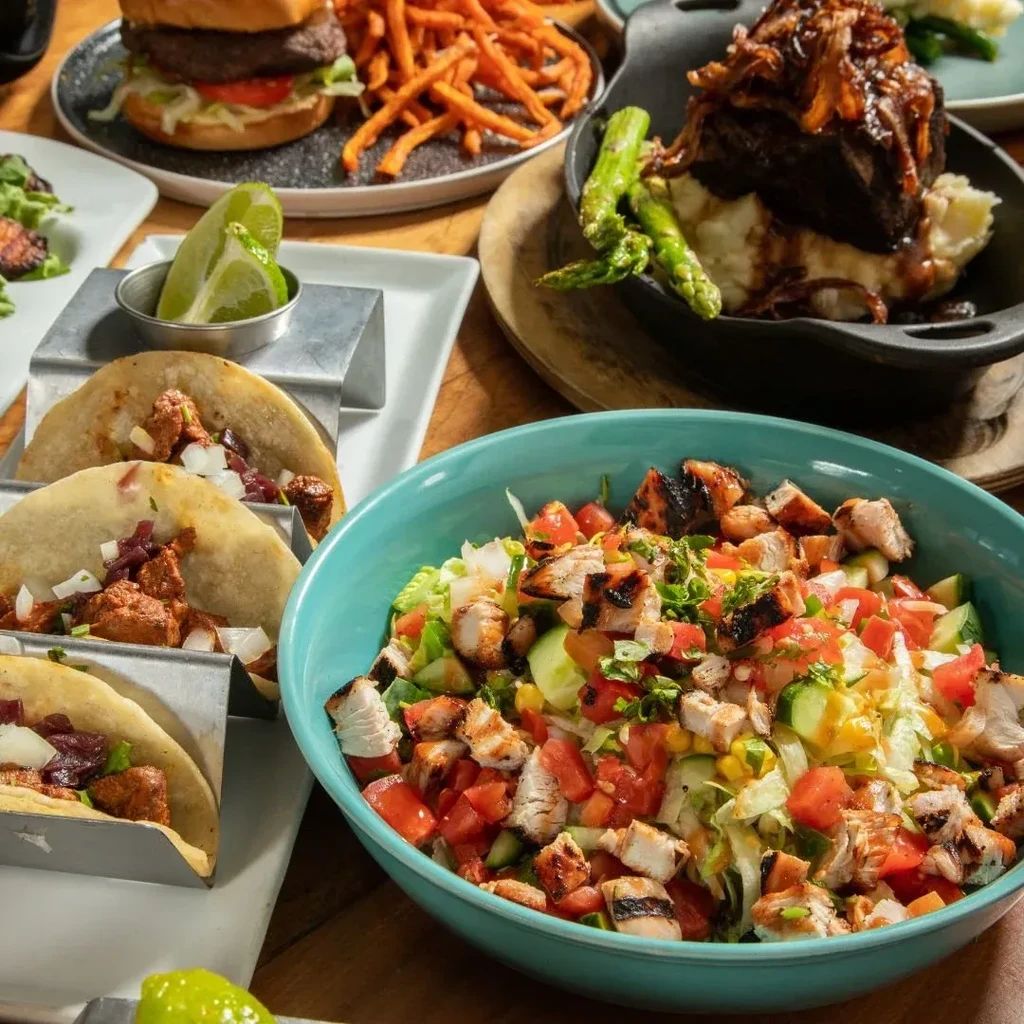 Overhead: chicken salad, tacos, burger, and braised meat on wooden table.