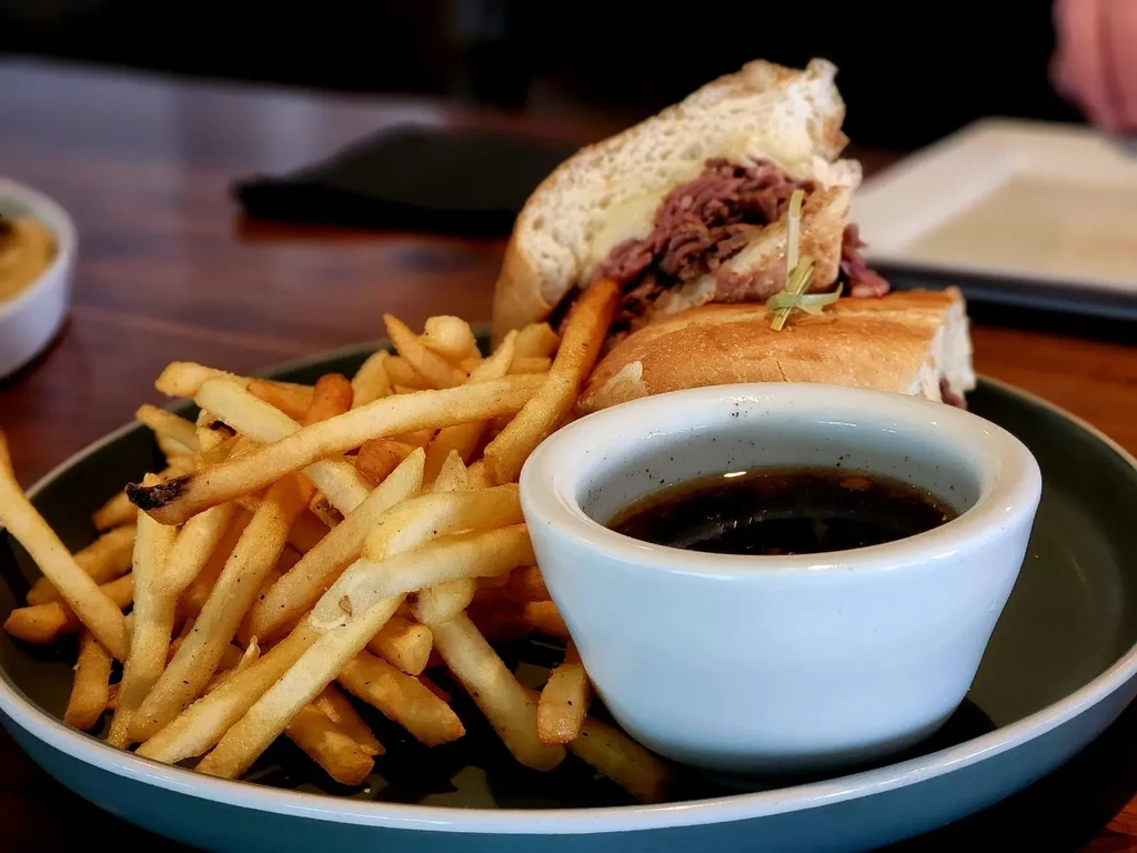 Large meat sandwich, fries, and sauce on a dark restaurant plate.
