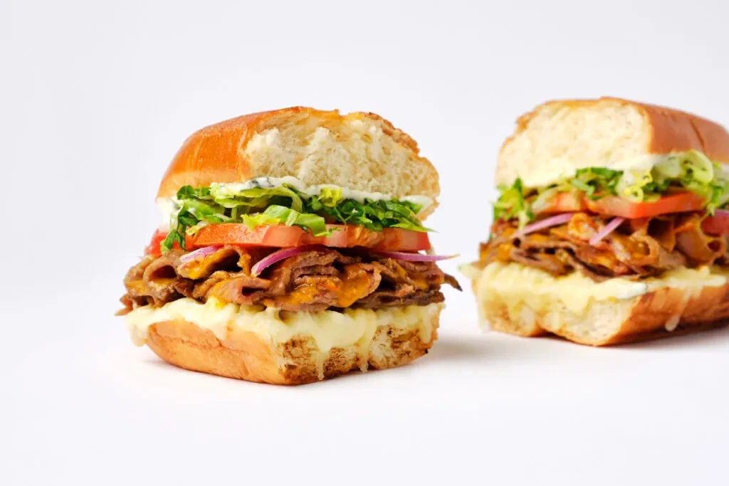 Two large sub sandwiches with meat, cheese, and vegetables on white.