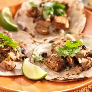 Three soft-shell tacos on orange plate with meat, rice, beans, cilantro, lime.