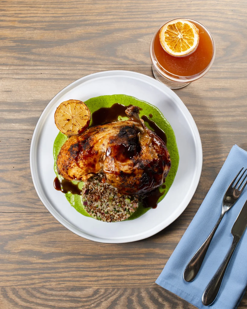 Glazed chicken on green purée, grains, cocktail, and napkin on wood.