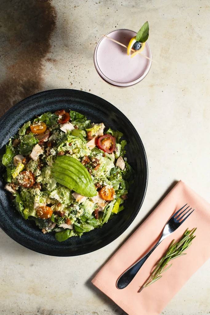 Overhead: chicken salad with avocado, pink cocktail, fork on stone.