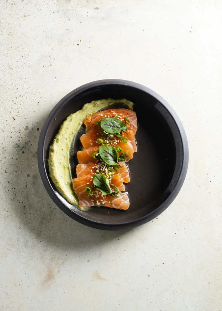 Top-down salmon sashimi with green sauce, herbs, and sesame seeds.