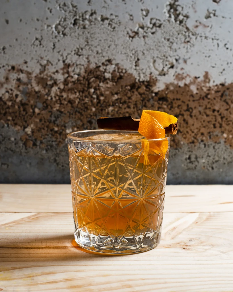 Amber cocktail in cut-glass tumbler with cinnamon stick and orange peel.