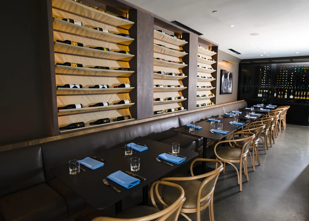 Contemporary restaurant with banquette, tables, and walls of wine bottles.