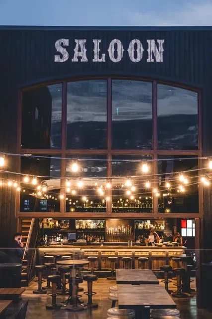 Modern saloon interior with lit bar, string lights, and high-top tables.