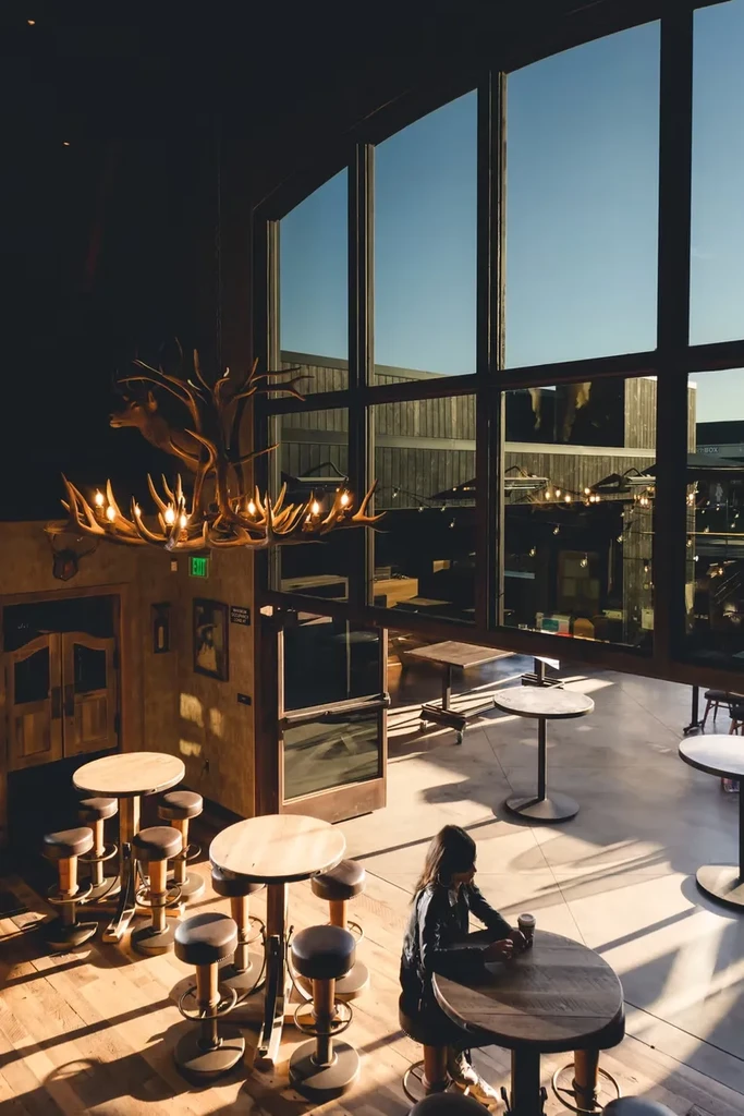 Lodge-style restaurant interior with antler chandelier, tall windows, one person.