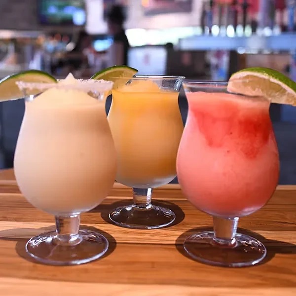 Three colorful frozen cocktails with lime wedges on a wooden bar.