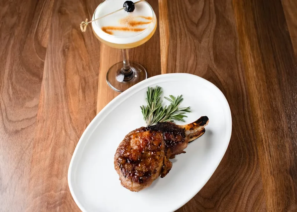Overhead view of a garnished pork chop and frothy cocktail.
