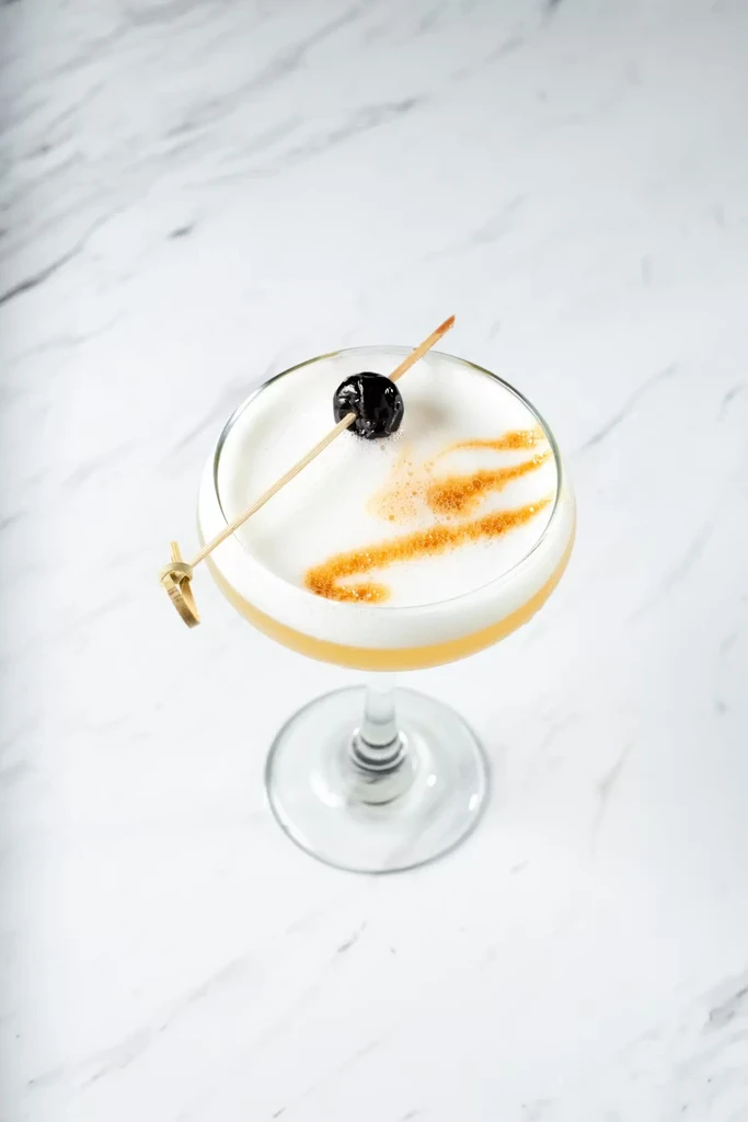 Cocktail in coupe glass with yellow liquid, white foam, cherry, golden drizzles.