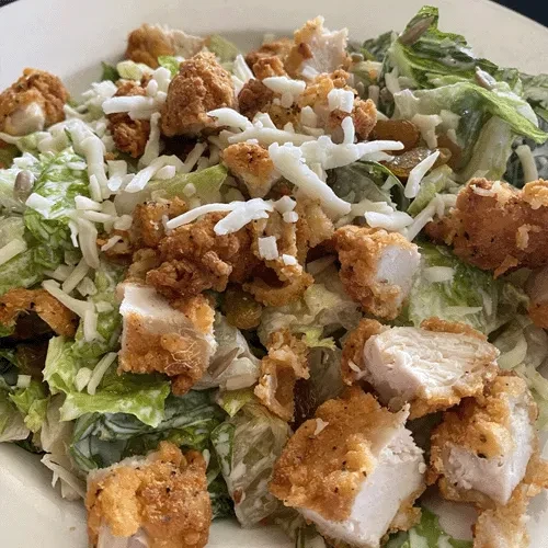 Salad in white bowl with crispy chicken, lettuce, cheese, raisins, seeds.