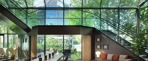 Modern glass lounge with staircase, sofa, communal table, and outdoor greenery.