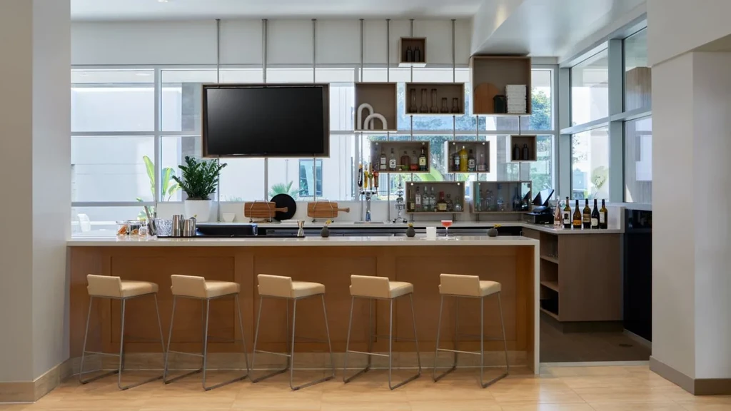 Modern bar with wood counter, five stools, liquor, and bright windows.