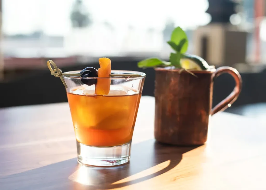 Amber cocktail and copper mug drink on a wooden table.