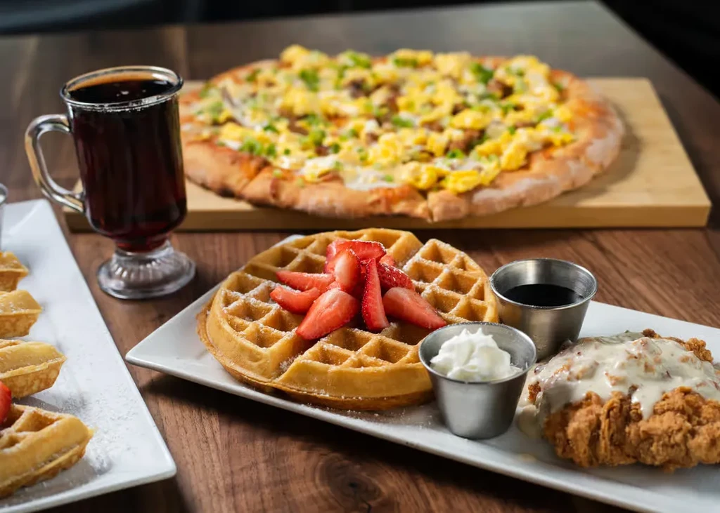 Waffle, fried chicken, and breakfast pizza on a dark table.