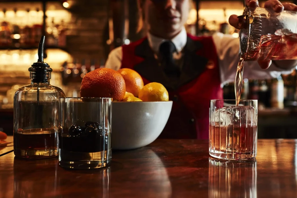 Bartender in red vest pouring cocktail into glass with ice.