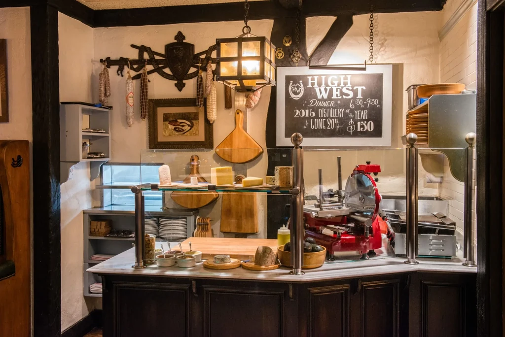 Rustic deli counter with cheese, meat slicer, hanging sausages, event chalkboard.