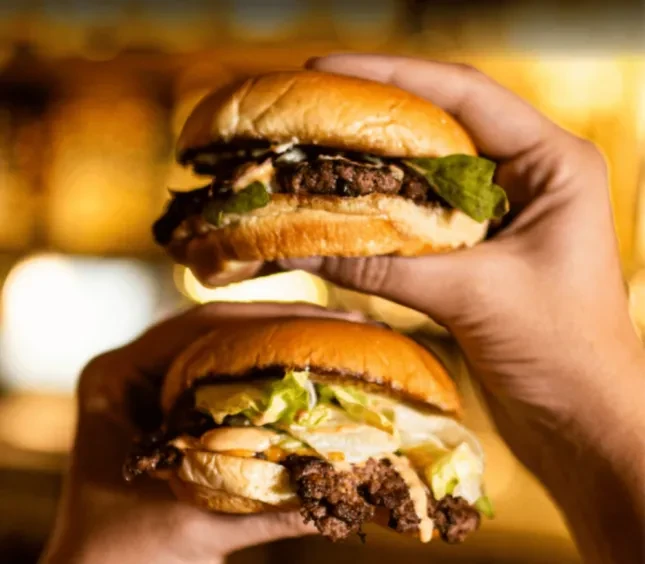Two hands hold two smash burgers with cheese, lettuce, and sauce.