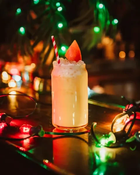 Yellow-orange drink with foam, fruit, straw, and blurred colorful lights.