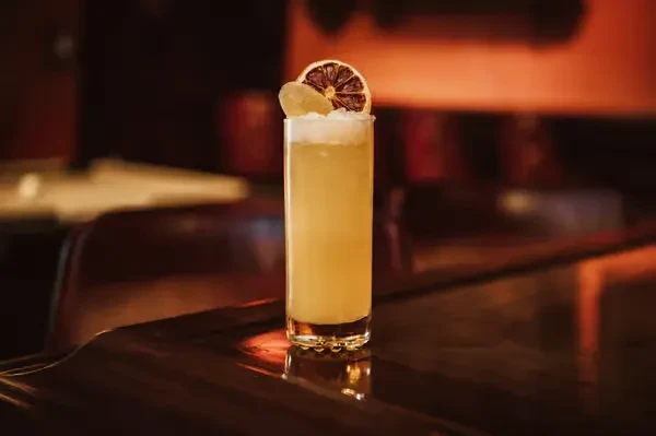 Garnished amber cocktail with a frothy layer on a dark bar.