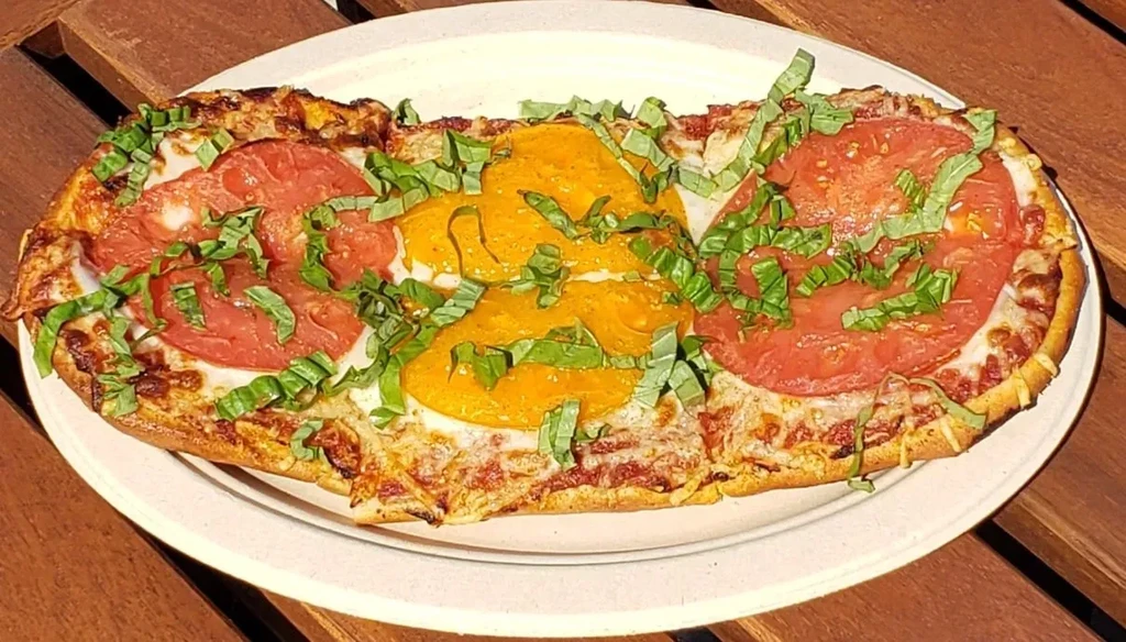 Rectangular flatbread pizza with red and yellow toppings, basil, and cheese.