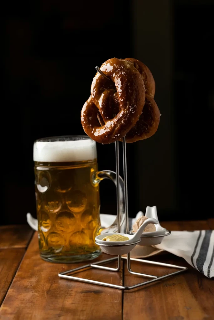 Two glazed salted pretzels on a stand with dips and beer.