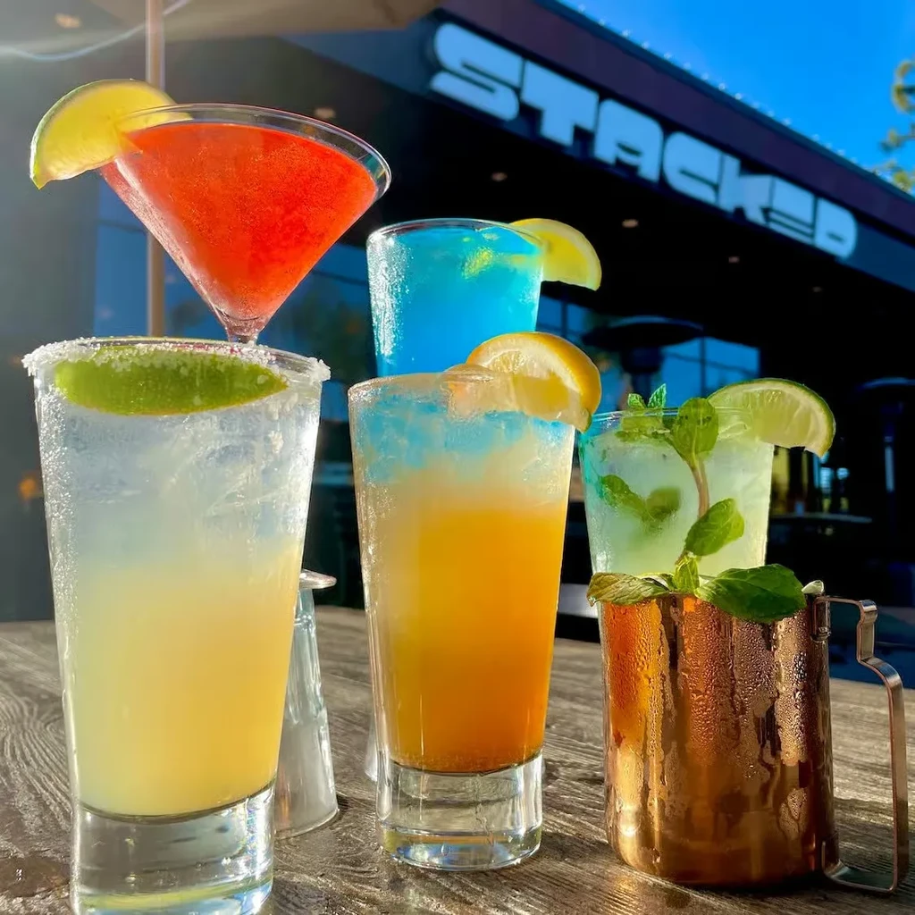 Six colorful cocktails on an outdoor table; "STACKED" sign in background.