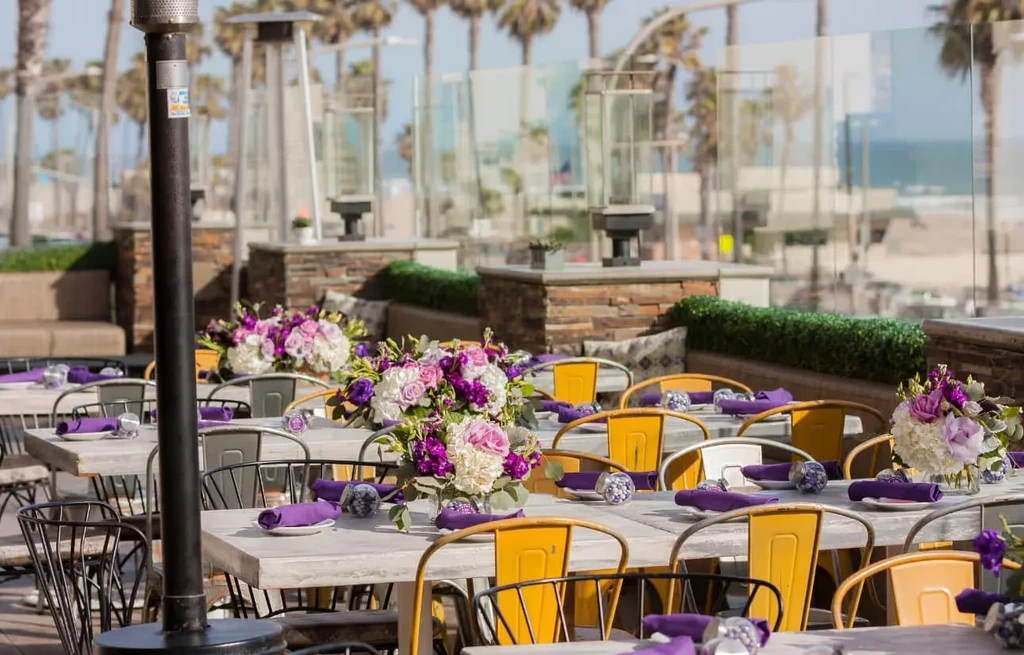 Restaurant patio tables set with purple flowers, yellow chairs, ocean view.