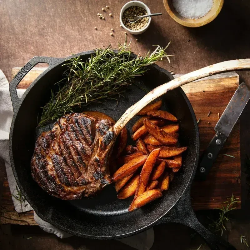 Grilled tomahawk steak, potato wedges, and rosemary in cast iron skillet.