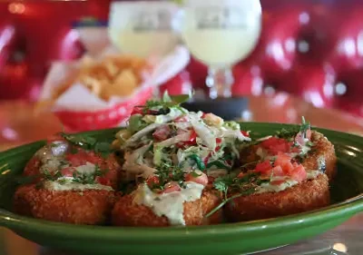 Fried food on green plate with sauce, tomatoes; drinks and chips in background.