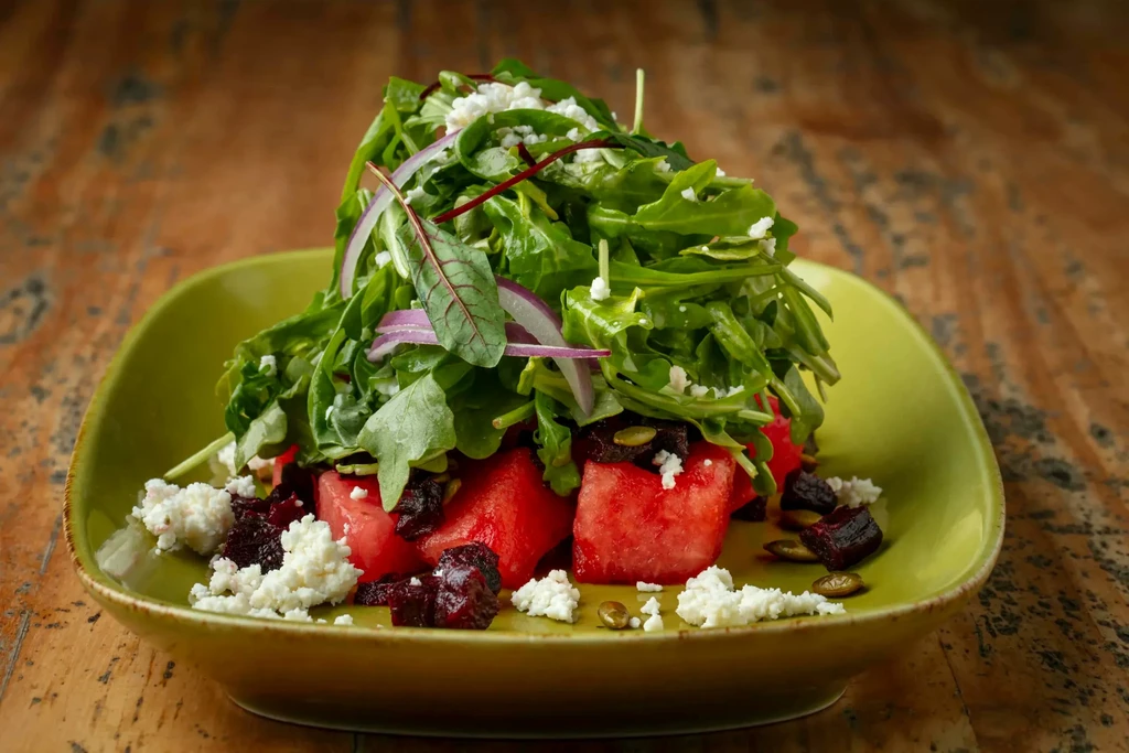 Arugula salad with watermelon, feta, cranberries, pumpkin seeds on green plate.