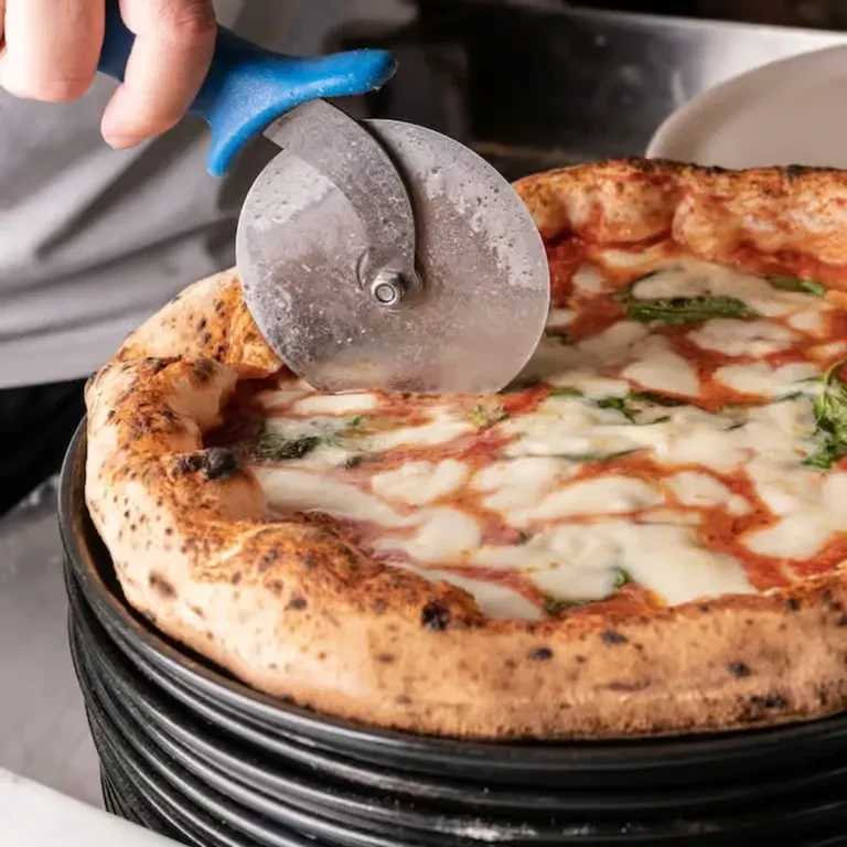 Hand slicing freshly baked pizza with mozzarella, tomato sauce, and basil.