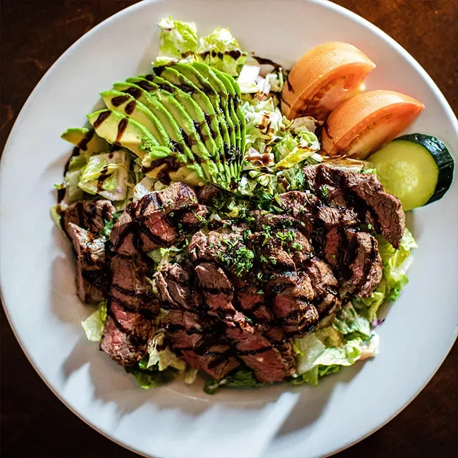 Grilled steak salad with avocado, tomato, and cucumber on a white plate.