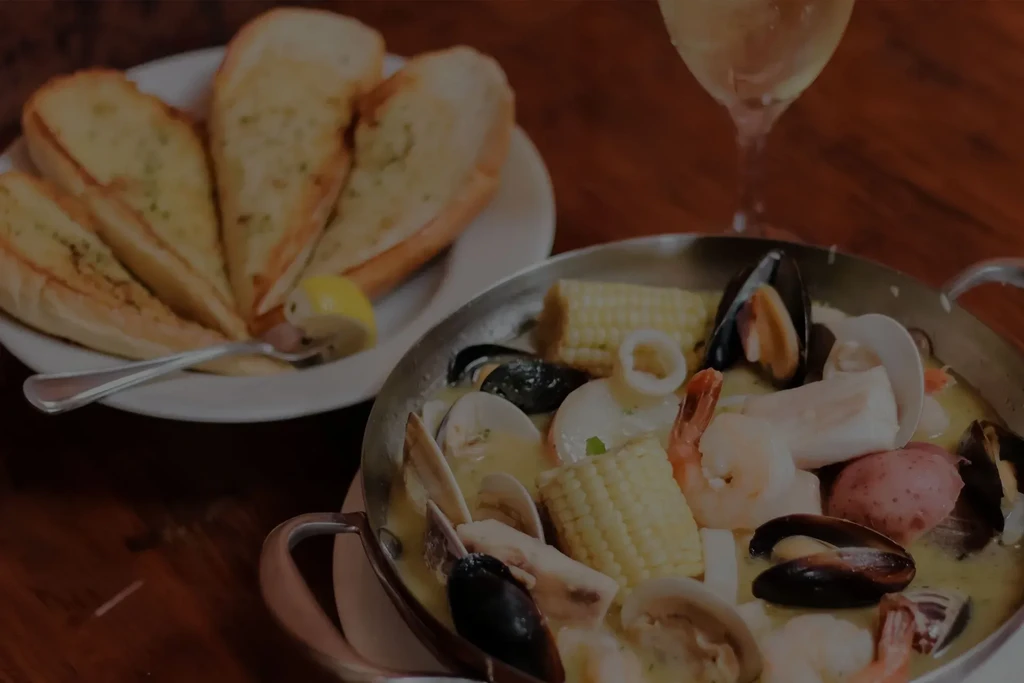 A pot of seafood stew, garlic bread, and wine on a table.