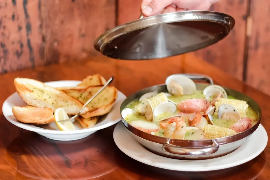 Hand lifting lid from steaming seafood boil, with garlic bread.