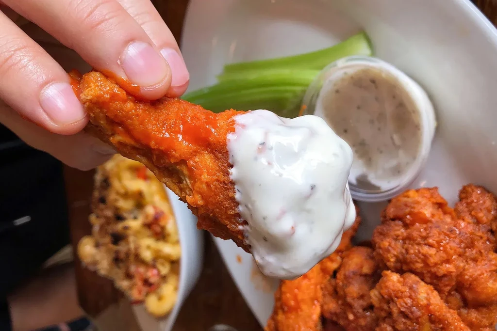 Hand holds buffalo chicken wing dripping creamy white sauce.