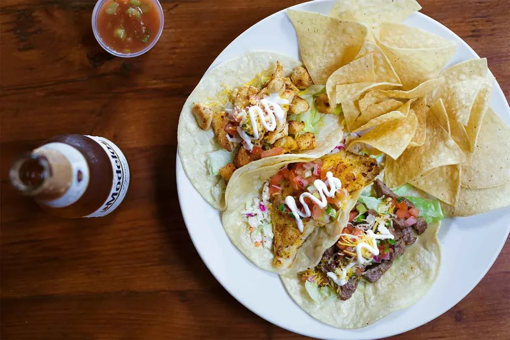 Three soft-shell tacos, chips, salsa, and Modelo beer on wooden table.