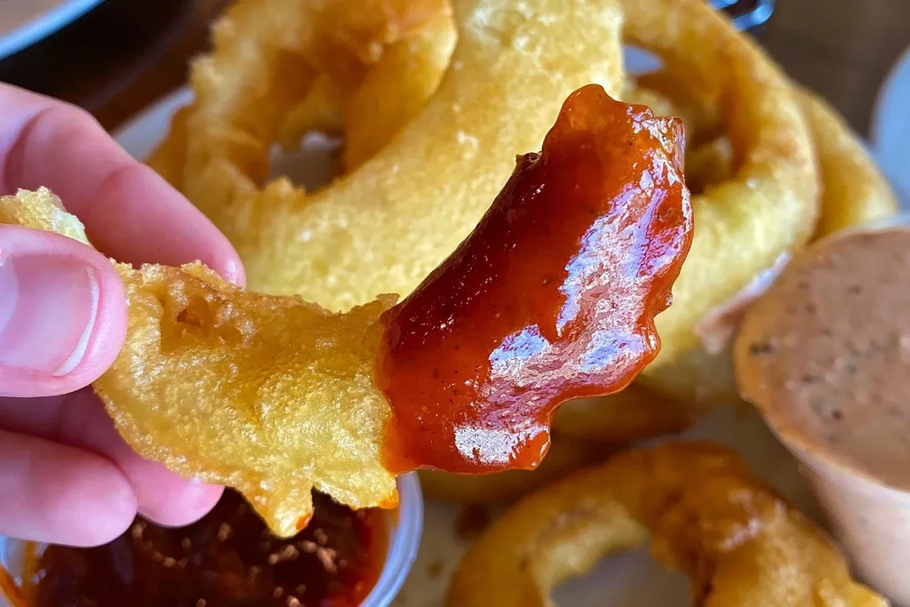 Hand dips a golden-brown onion ring into red sauce, more rings visible.