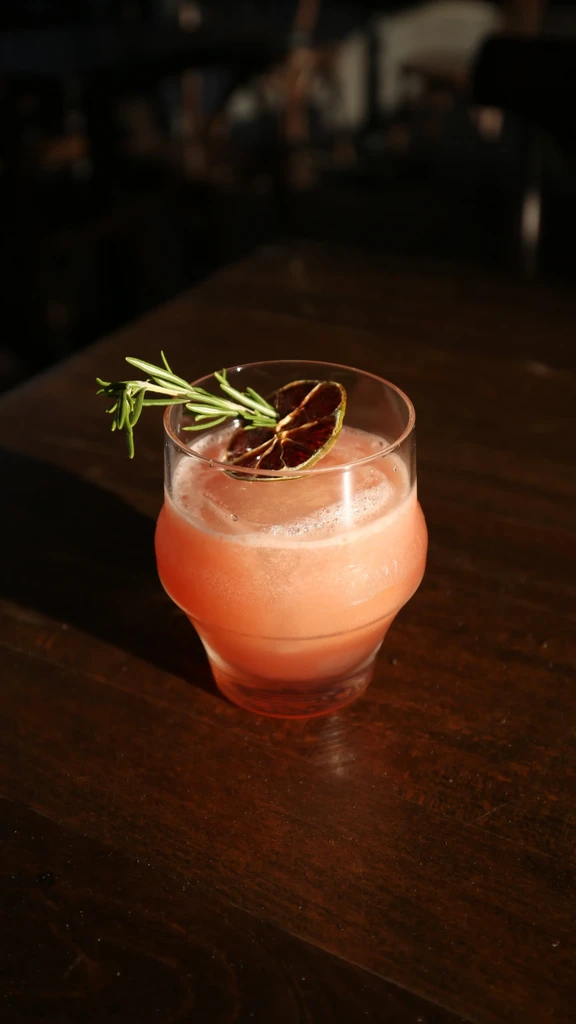 Reddish-orange foamy cocktail in textured glass with rosemary and dried citrus.