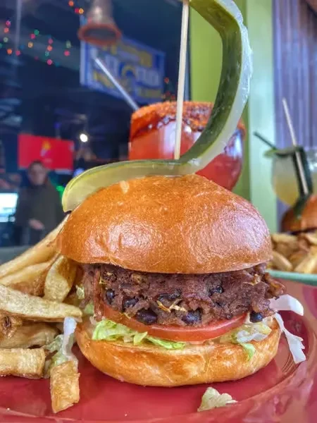 Gourmet burger with dark patty, fries, and a skewered pickle.